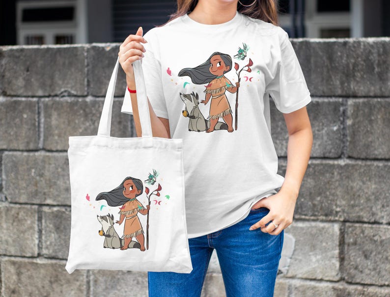 May include: White tote bag with a cartoon illustration of a Native American girl with long black hair, wearing a brown dress and holding a staff. She is accompanied by a raccoon. The bag has a white background with pink and blue floral accents.