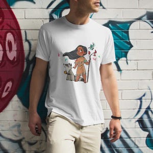 May include: White t-shirt with a cartoon image of a Native American girl with long black hair, wearing a brown and white dress, holding a stick with a blue bird on it. A raccoon is standing next to her.