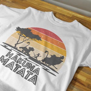 May include: White t-shirt featuring a retro sunset design with silhouettes of characters from a popular animated film. The phrase "HAKUNA MATATA" is printed in bold, stylized letters below the graphic.