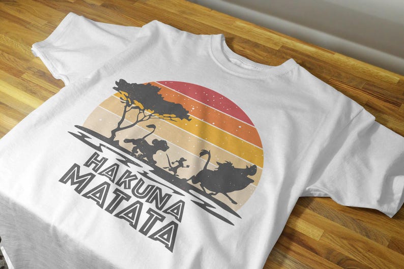May include: White t-shirt featuring a retro sunset design with silhouettes of characters from a popular animated film. The phrase "HAKUNA MATATA" is printed in bold, stylized letters below the graphic.