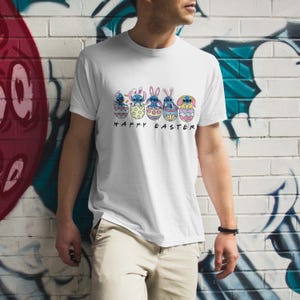 May include: White t-shirt with a graphic of five blue Stitch characters from the Disney movie Lilo & Stitch, each inside a colorful Easter egg. The text "Happy Easter" is printed below the eggs.