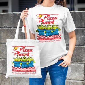 May include: White tote bag with a green alien character design. The bag has the text "Pizza Planet" and "Food & Fun Space Port" printed on it. The bag is being held by a person wearing a white t-shirt with the same design.