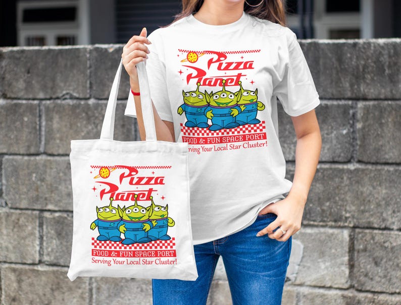 May include: White tote bag with a green alien character design. The bag has the text "Pizza Planet" and "Food & Fun Space Port" printed on it. The bag is being held by a person wearing a white t-shirt with the same design.