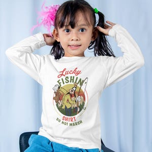May include: A white long-sleeved shirt with a cartoon character of Goofy fishing. The shirt has a vintage-style graphic with the text "Lucky Fishin' Shirt Do Not Wash".