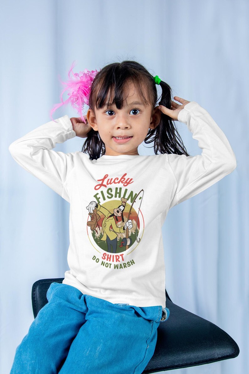 May include: A white long-sleeved shirt with a cartoon character of Goofy fishing. The shirt has a vintage-style graphic with the text "Lucky Fishin' Shirt Do Not Wash".