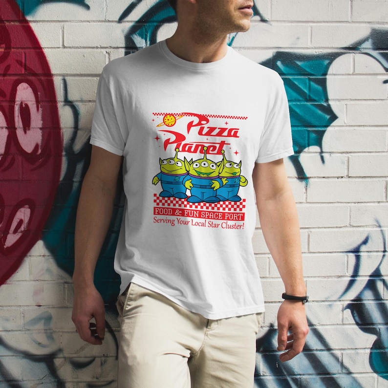 May include: White t-shirt with a graphic of the Pizza Planet restaurant from the Toy Story movies. The graphic features three green aliens with the text "Pizza Planet" and "Food & Fun Space Port" in red. The text "Serving Your Local Star Cluster!" is also included in red.