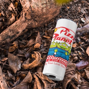 May include: White travel mug with a red, white, and blue design featuring the words "Pizza Planet" and two green alien characters from the Toy Story movie franchise. The mug is sitting on a bed of brown leaves.
