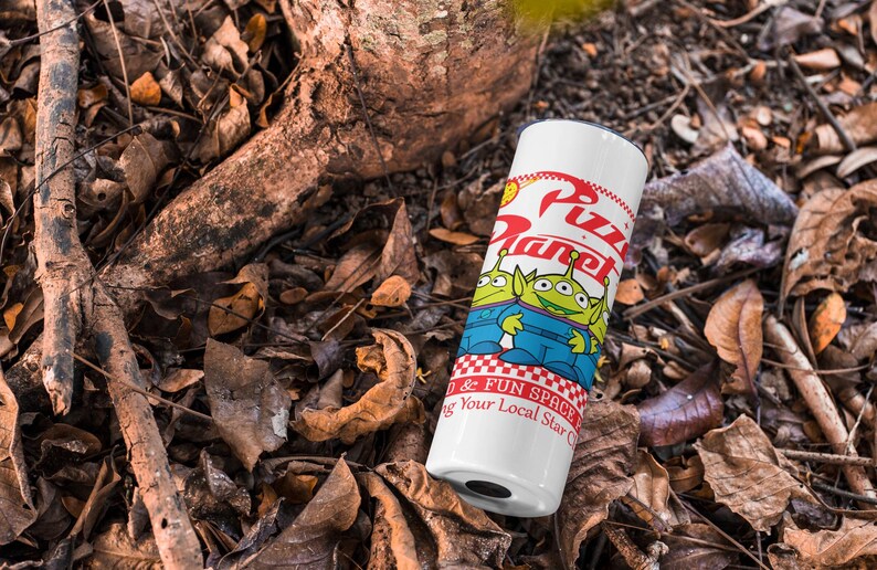 May include: White travel mug with a red, white, and blue design featuring the words "Pizza Planet" and two green alien characters from the Toy Story movie franchise. The mug is sitting on a bed of brown leaves.