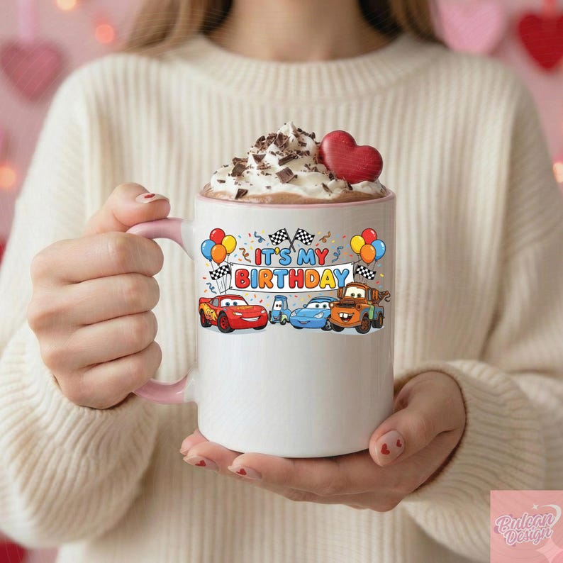 May include: White ceramic mug with a pink handle and rim, featuring a colorful "It's My Birthday" design with cartoon cars, balloons, and checkered flags. The mug is filled with whipped cream, chocolate shavings, and a red heart.