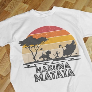 May include: White t-shirt featuring a retro sunset design with silhouettes of characters from The Lion King. The phrase "Hakuna Matata" is printed in bold, black letters below the image. The shirt is made of a soft material.