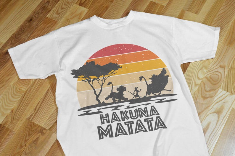 May include: White t-shirt featuring a retro sunset design with silhouettes of characters from The Lion King. The phrase "Hakuna Matata" is printed in bold, black letters below the image. The shirt is made of a soft material.