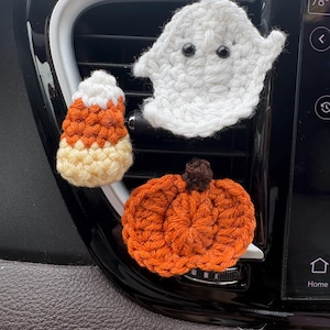 May include: Three crocheted Halloween decorations attached to a car air vent. The set includes a white ghost with black bead eyes, a candy corn with white, orange, and yellow sections, and a round orange pumpkin with a small brown stem on top.
