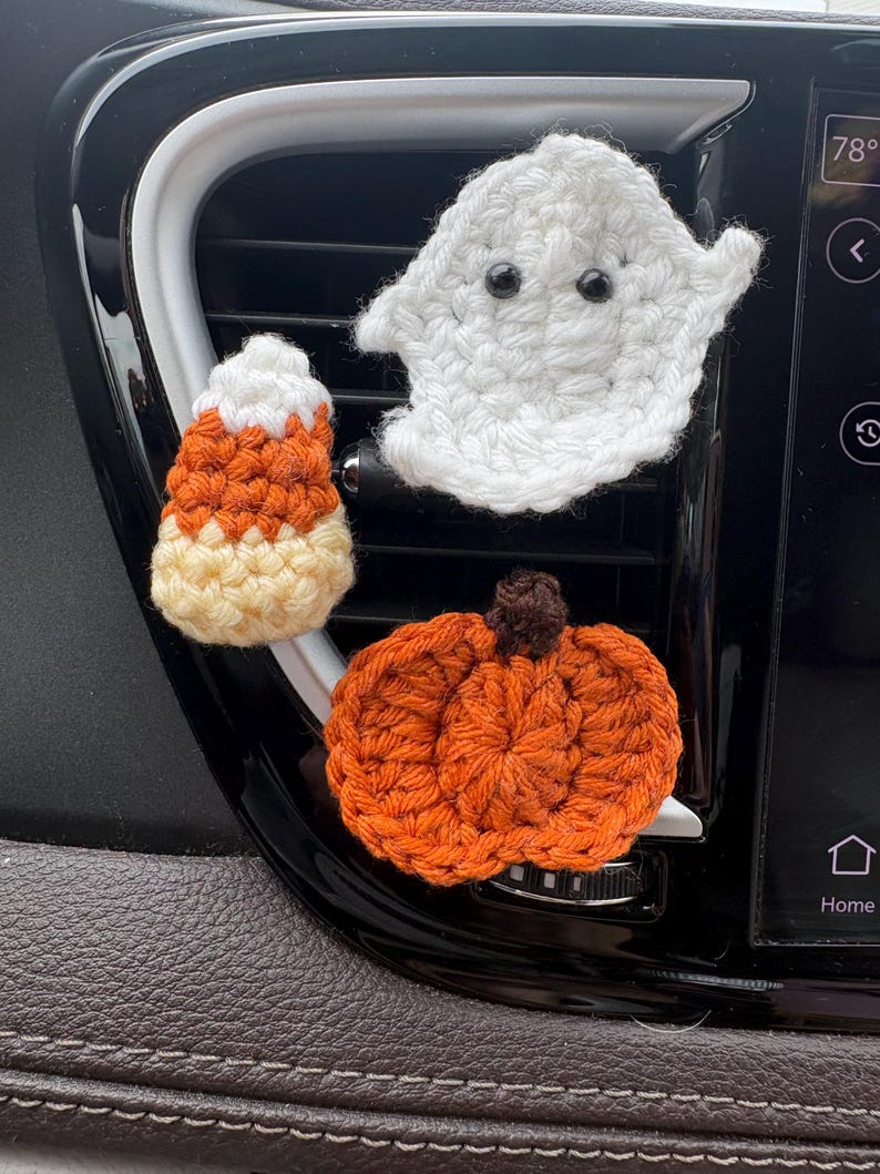 Puede incluir: Tres decoraciones de Halloween tejidas a ganchillo sujetas a la rejilla de ventilaci&oacute;n de un coche. El conjunto incluye un fantasma blanco con ojos de cuentas negras, un peque&ntilde;o caramelo de ma&iacute;z naranja, blanco y amarillo, y una calabaza.
