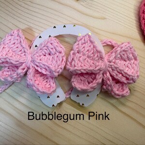 May include: A close-up of three bubblegum pink crochet bows attached to a white horseshoe-shaped base with gold heart accents. The text "Bubblegum Pink" is displayed below the bows. A pink crochet heart is visible in the background.