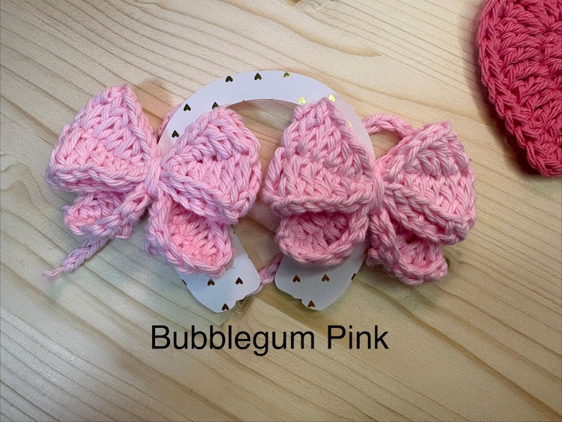 May include: A close-up of three bubblegum pink crochet bows attached to a white horseshoe-shaped base with gold heart accents. The text "Bubblegum Pink" is displayed below the bows. A pink crochet heart is visible in the background.
