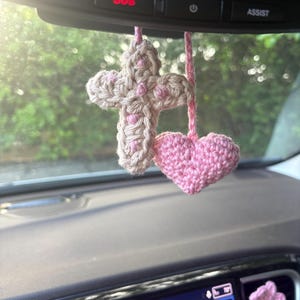May include: A beige crocheted cross with pink accents and a solid pink crocheted heart hanging from a rearview mirror. A small pink crocheted flower is attached to the dashboard console, which displays a digital screen with the text SOS and ASSIST.
