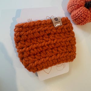 May include: A burnt orange crochet cup sleeve made of thick yarn, featuring a small rectangular wooden tag labelled "handmade" with a yarn ball icon. Next to it is a small, round, crocheted orange pumpkin with a dark brown stem.