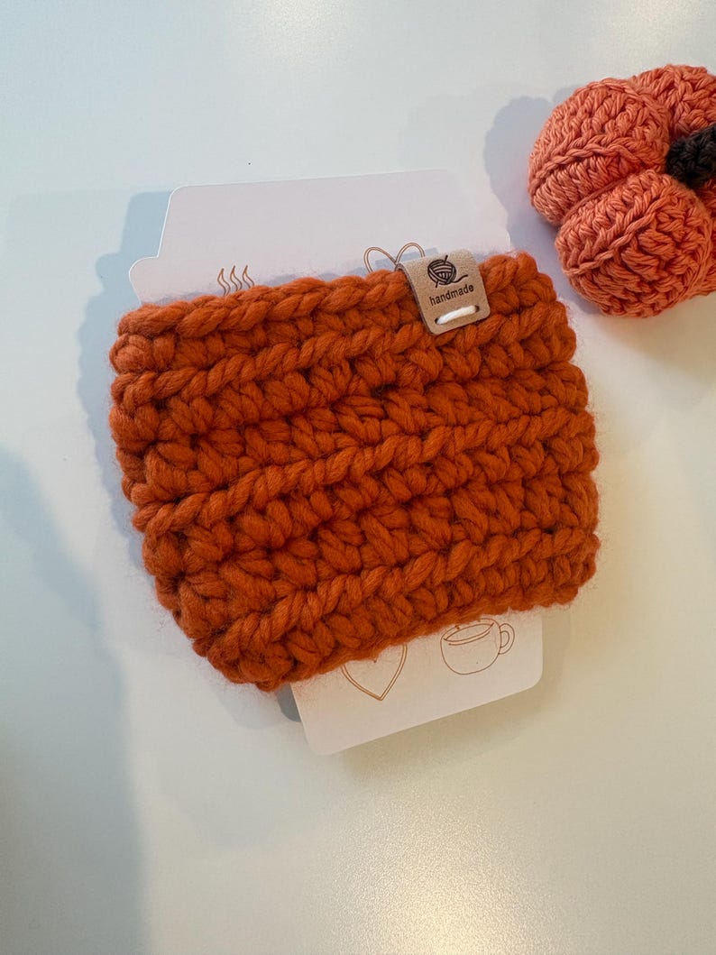 May include: A burnt orange crochet cup sleeve made of thick yarn, featuring a small rectangular wooden tag labelled "handmade" with a yarn ball icon. Next to it is a small, round, crocheted orange pumpkin with a dark brown stem.