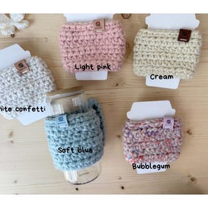 May include: Five crochet cup sleeves in white confetti, light pink, cream, soft blue, and bubblegum colours. Each sleeve features a small rectangular tag and is displayed on a white card. One soft blue sleeve is wrapped around a glass tumbler.