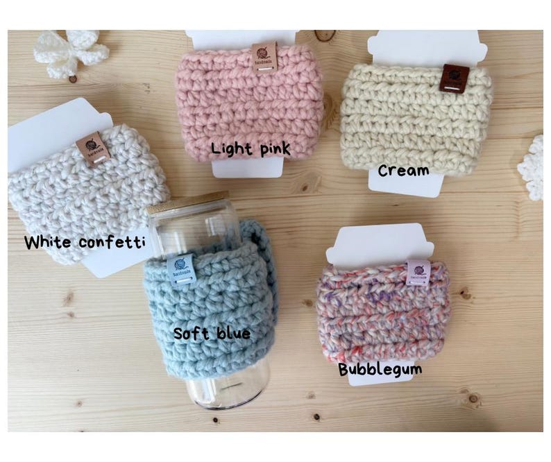 May include: Five crochet cup sleeves in white confetti, light pink, cream, soft blue, and bubblegum colours. Each sleeve features a small rectangular tag and is displayed on a white card. One soft blue sleeve is wrapped around a glass tumbler.