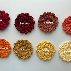May include: Eight circular crochet flower appliques arranged in two rows. The colors are labeled as Red, Burnt Sienna, Bruschetta, Orange, Glowing Orange, Gold, Sunshine, and Buttercup. Each piece features a textured, puff-stitch petal design.