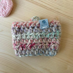 May include: A crochet cup sleeve made of variegated yarn in shades of pink, blue, green, and cream. The sleeve features a small, rectangular, light blue tag with a graphic of a ball of yarn and the word handmade printed in dark blue text.