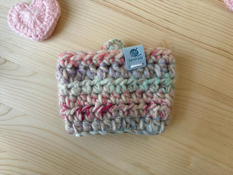 May include: A crochet cup sleeve made of variegated yarn in shades of pink, blue, green, and cream. The sleeve features a small, rectangular, light blue tag with a graphic of a ball of yarn and the word handmade printed in dark blue text.