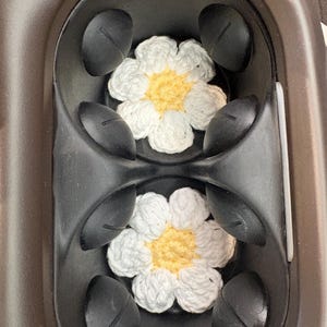 May include: Two white and yellow crocheted daisy coasters are placed inside a black car cup holder. The coasters have white petals and yellow centers. The cup holder is black and brown.