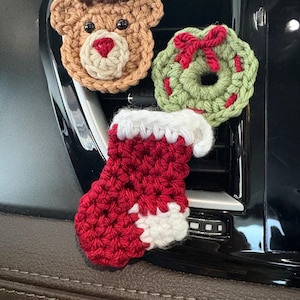 Puede incluir: Tres adornos navideños de ganchillo que incluyen un reno color canela con astas marrón oscuro, una corona verde con un lazo rojo y un calcetín rojo con puño y punta blancos. Estas pequeñas decoraciones de hilo texturizado están fijadas sobre una superficie negra.