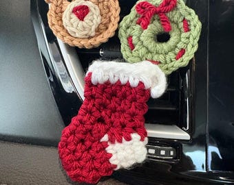 May include: Three crocheted Christmas ornaments featuring a tan reindeer with dark brown antlers, a green wreath with a red bow, and a red stocking with a white cuff and toe. These small, textured yarn decorations are attached to a black surface.