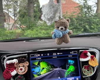 May include: A crochet car accessory set featuring a hanging brown bear with a blue scarf and a white snowflake. The dashboard vents are decorated with crochet pieces including a red stocking, a bear face, holly, a Santa hat, a bell, and a wreath.