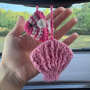 May include: A hanging car accessory featuring a small, crocheted pink and white striped fish with a black eye and a separate crocheted light pink seashell, both suspended from pink yarn strings attached to a rearview mirror.