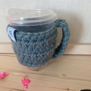 May include: A clear plastic cup with a lid, encased in a thick, chunky, slate-blue crocheted yarn cosy that features a matching handle and a small blue fabric tag attached to the side with a white graphic icon of a yarn ball.