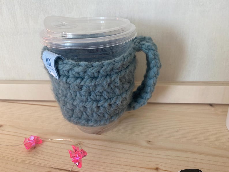 May include: A clear plastic cup with a lid, encased in a thick, chunky, slate-blue crocheted yarn cosy that features a matching handle and a small blue fabric tag attached to the side with a white graphic icon of a yarn ball.