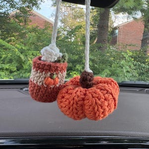 May include: A crochet car accessory featuring a small mug with a white whipped topping and a tiny orange pumpkin, paired with a larger orange pumpkin. Both items are suspended by white braided yarn strings from a rearview mirror mount.