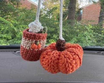May include: A crochet car hanging ornament featuring a small orange pumpkin and a mug with a white whipped topping, a tiny pumpkin accent, and a green leaf, both suspended by white braided cords from a rearview mirror mount.