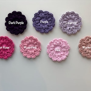 May include: Seven circular crochet flower appliques arranged in two rows. The top row features Dark Purple, Purple, and Lavender. The bottom row includes Hot Rose, Rose Pink, Pink, and Warm Blush. Each piece has a textured, puff-stitch design.