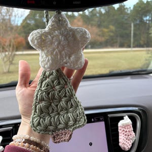 Puede incluir: Un accesorio colgante para coche que presenta una estrella de ganchillo blanca sobre un árbol de Navidad de ganchillo verde salvia con un tronco marrón. Un pequeño calcetín de ganchillo rosa está sujeto a la rejilla de ventilación.