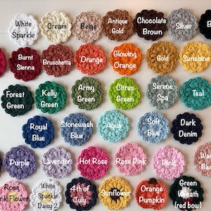 May include: A collection of small, round, crocheted flower appliques arranged in rows. Each piece features a textured, petal-like design and is labeled with its specific color name, including White Sparkle, Cream, Beige, Antique Gold, Chocolate Brown, Silver, Black, Red, Burnt Sienna, Bruschetta, Orange, Glowing Orange, Gold, Sunshine, Buttercup, Forest Green, Kelly Green, Army Green, Citrus Green, Serene Spa, Teal, Royal Blue, Stonewash Blue, Aqua, Soft Blue, Dark Denim, Dark Purple, Purple, Lavender, Hot Rose, Rose Pink, Pink, Warm Blush, Rose Pink Flower, White Sparkle Daisy 2, 4th of July, Sunflower, Orange Pumpkin, and Green Wreath with red bow.