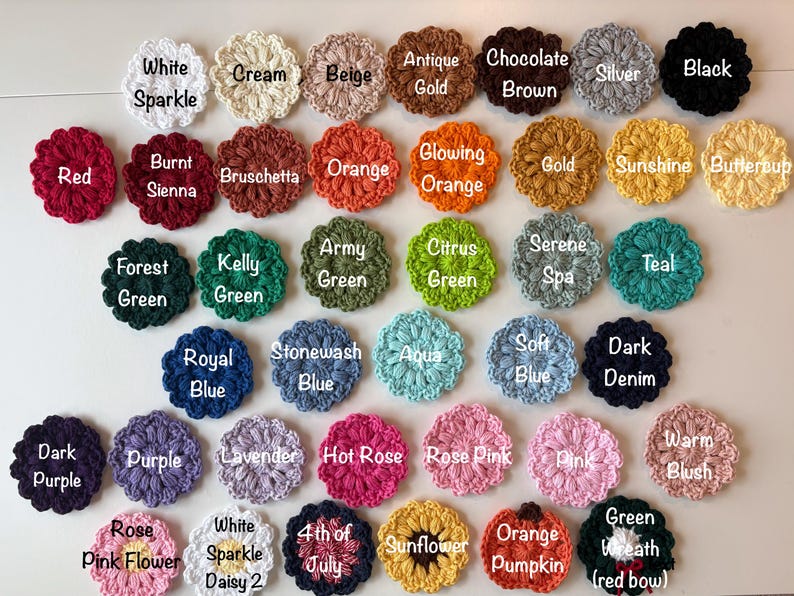 May include: A collection of small, round, crocheted flower appliques arranged in rows. Each piece features a textured, petal-like design and is labeled with its specific color name, including White Sparkle, Cream, Beige, Antique Gold, Chocolate Brown, Silver, Black, Red, Burnt Sienna, Bruschetta, Orange, Glowing Orange, Gold, Sunshine, Buttercup, Forest Green, Kelly Green, Army Green, Citrus Green, Serene Spa, Teal, Royal Blue, Stonewash Blue, Aqua, Soft Blue, Dark Denim, Dark Purple, Purple, Lavender, Hot Rose, Rose Pink, Pink, Warm Blush, Rose Pink Flower, White Sparkle Daisy 2, 4th of July, Sunflower, Orange Pumpkin, and Green Wreath with red bow.