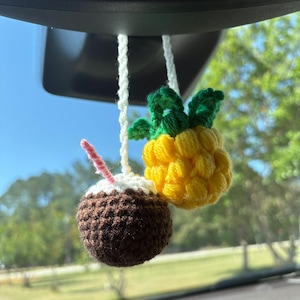 May include: Crocheted car charm featuring a brown coconut drink with a pink straw and a yellow pineapple with green leaves. The handmade ornaments hang from a white string, adding a tropical touch to the car's interior.