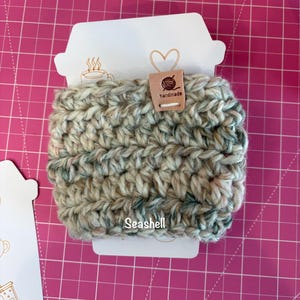 May include: A crochet cup sleeve in a variegated yarn featuring shades of cream, tan, and muted green. The sleeve is attached to a white cardboard display card with a coffee cup shape, featuring the text "Seashell" and a small "handmade" tag.