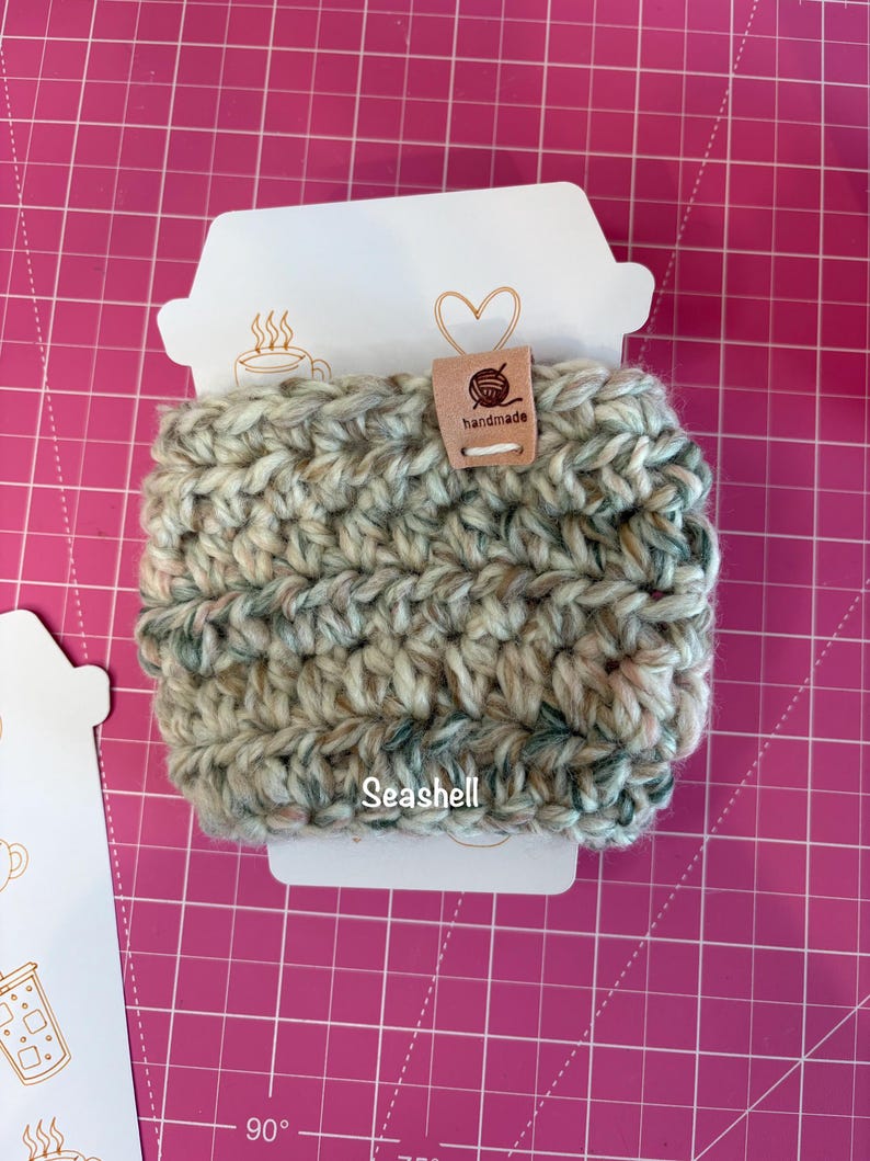 May include: A crochet cup sleeve in a variegated yarn featuring shades of cream, tan, and muted green. The sleeve is attached to a white cardboard display card with a coffee cup shape, featuring the text "Seashell" and a small "handmade" tag.