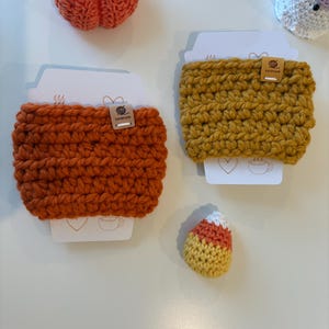 May include: Two crochet cup sleeves in orange and mustard yellow, each featuring a small handmade tag. The sleeves are displayed on white cards with line art icons. Nearby are a small crocheted orange pumpkin, a white ghost, and candy corn.
