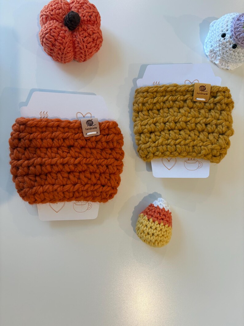 May include: Two crochet cup sleeves in orange and mustard yellow, each featuring a small handmade tag. The sleeves are displayed on white cards with line art icons. Nearby are a small crocheted orange pumpkin, a white ghost, and candy corn.
