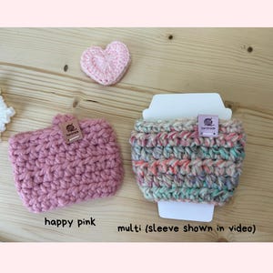 May include: Two crocheted cup sleeves, one solid pink and one multicoloured, each featuring a small rectangular tag labelled handmade. A small pink crocheted heart is positioned above the pink sleeve. Text below reads happy pink and multi (sleeve shown in video).