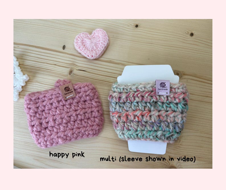 May include: Two crocheted cup sleeves, one solid pink and one multicoloured, each featuring a small rectangular tag labelled handmade. A small pink crocheted heart is positioned above the pink sleeve. Text below reads happy pink and multi (sleeve shown in video).
