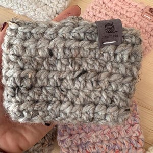 May include: A grey, chunky knit crochet cup sleeve featuring a dark grey fabric tag with a yarn ball icon and the text "handmade" stitched in white. The sleeve is crafted from thick, textured yarn with subtle dark flecks throughout the material.
