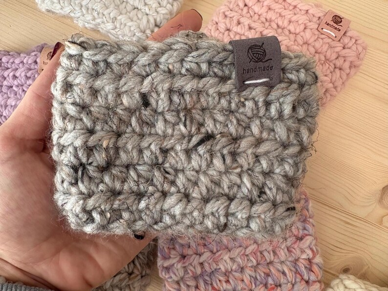 May include: A grey, chunky knit crochet cup sleeve featuring a dark grey fabric tag with a yarn ball icon and the text "handmade" stitched in white. The sleeve is crafted from thick, textured yarn with subtle dark flecks throughout the material.