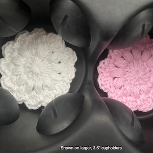 May include: Two circular crochet coasters, one white and one light pink, featuring a textured floral pattern. These coasters are positioned inside a black plastic cup holder assembly, with text at the bottom stating shown on larger, 3.5" cupholders.
